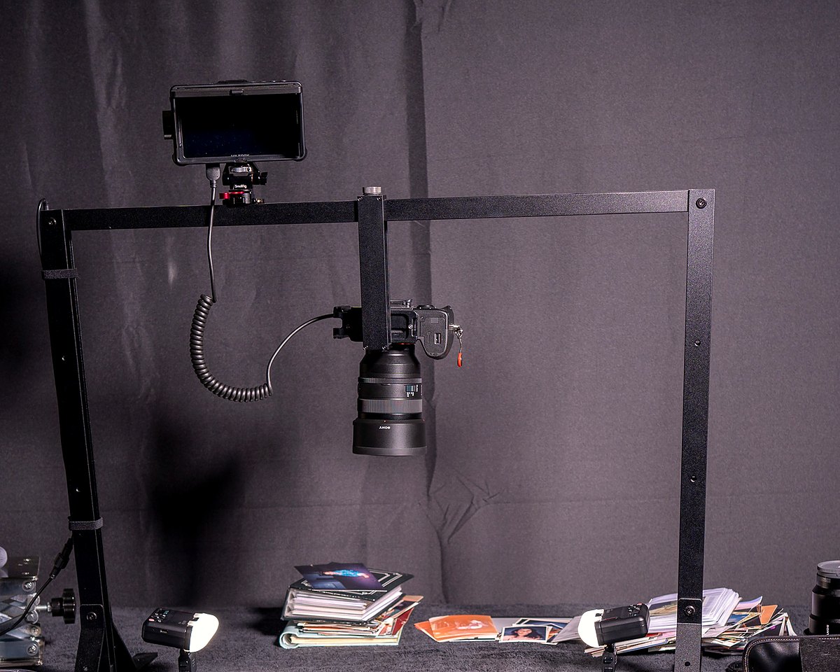 The studio copy stand: a camera mounted overhead with a monitor, two small lights either side and a stack of family prints on the table below